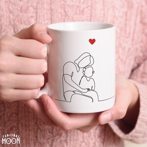 May include: White ceramic mug with a black line drawing of a parent holding a child. A small red heart is at the top of the mug.