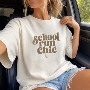 May include: A cream-colored short-sleeve t-shirt featuring the brown text "school run Chic" in a wavy, retro-inspired font. Below the text is a small brown crescent moon and star graphic. The shirt has a crew neckline and a relaxed fit.