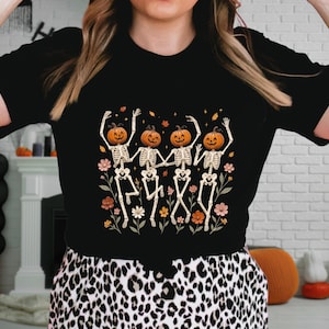 May include: A black short-sleeved t-shirt featuring a graphic print of four skeletons with orange pumpkin heads dancing in a line. The skeletons are surrounded by colorful autumn-themed flowers and small leaves on a dark background.