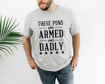 May include: A heather gray short-sleeved t-shirt featuring black text that reads "THESE PUNS ARE ARMED AND DADLY" with five small black stars underneath the lettering. The shirt has a crew neckline and a standard fit, displaying the graphic on the chest.