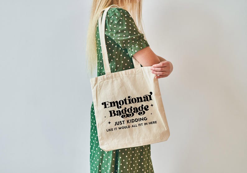May include: A beige canvas tote bag featuring black text that reads Emotional Baggage, JUST KIDDING, LIKE IT WOULD ALL FIT IN HERE, accented with small four-pointed stars. The bag has two long shoulder straps and a rectangular, flat-bottomed design.