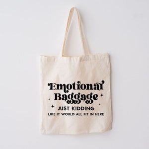 May include: A beige canvas tote bag with two shoulder straps. The front features black text that reads Emotional Baggage, JUST KIDDING, LIKE IT WOULD ALL FIT IN HERE, accented by four small four-pointed star symbols surrounding the main text.