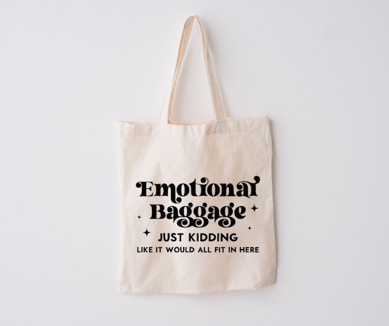 May include: A beige canvas tote bag with two shoulder straps. The front features black text that reads Emotional Baggage, JUST KIDDING, LIKE IT WOULD ALL FIT IN HERE, accented by four small four-pointed star symbols surrounding the main text.