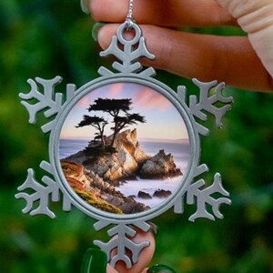 May include: A silver snowflake ornament with a photo of a lone tree on a rocky coastline. The photo shows a sunset sky with pink and orange hues.