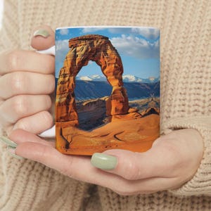 May include: A white ceramic mug with a colorful image of Delicate Arch in Arches National Park, Utah. The arch is a natural sandstone formation with a blue sky and clouds in the background.