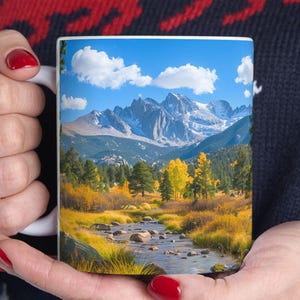 May include: A white ceramic mug with a scenic image of a mountain range with snow-capped peaks, a river flowing through a valley, and fall foliage in shades of yellow, orange, and red.