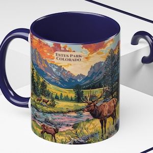 May include: A blue and white ceramic mug with a scenic image of a mountain range, a river, and elk. The mug has the text "Estes Park Colorado" printed on it.