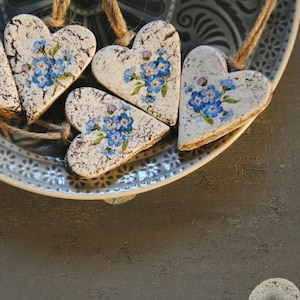 Könnte beinhalten: Fünf herzförmige Ornamente mit einem weißen Used-Look, verziert mit blauen Blumenmustern, liegen auf einem dekorativen blau-weißen Teller. Die Ornamente sind mit Schnur verbunden. Der Hintergrund ist eine dunkelgraue Oberfläche.