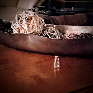 Può includere: Una ciotola d'argento piena di sfere decorative intrecciate e una piccola bottiglia di vetro con un tappo di sughero. La ciotola è appoggiata su una superficie di legno.