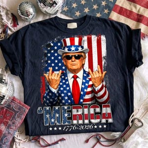Peut inclure: Un t-shirt bleu marine foncé avec un motif représentant une personne en costume étoilé rouge, blanc et bleu, haut-de-forme et lunettes de soleil, faisant un geste de la main rock. Le texte indique 'MERICA 1776-2026' en police grasse et usée.
