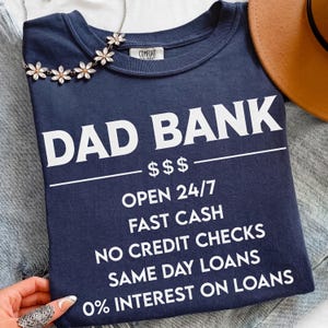 May include: Navy blue t-shirt with the text "DAD BANK" in large white letters. The shirt also features the phrases "OPEN 24/7", "FAST CASH", "NO CREDIT CHECKS", "SAME DAY LOANS", and "0% INTEREST ON LOANS". A floral necklace is draped over the shirt.