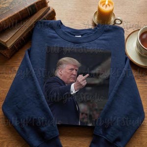 May include: Navy blue sweatshirt featuring a photographic print of a man in a suit, pointing his middle finger. The image is centered on the front of the sweatshirt. The sweatshirt is displayed on a wooden surface with books and a candle.