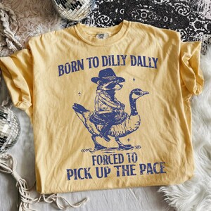 May include: Yellow t-shirt with a blue graphic of a raccoon in a cowboy hat riding a goose. The text "BORN TO DILLY DALLY" is above the raccoon, and "FORCED TO PICK UP THE PACE" is below.