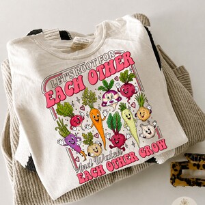 Puede incluir: Una camiseta de color crema con un estampado gráfico de varias verduras de dibujos animados con caras. El texto dice "LET'S ROOT FOR EACH OTHER" y "AND WATCH EACH OTHER GROW" en letras rosas. El diseño incluye una zanahoria, un rábano y una cebolla.