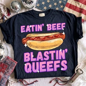 May include: A black short-sleeve t-shirt featuring a graphic of a hot dog with red ketchup. The text EATIN' BEEF is printed in pink above the hot dog, and BLASTIN' QUEEFS is printed in pink below, accented with small white star sparkles.