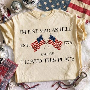 Może przedstawiać: Kremowa koszulka z napisem "I'M JUST MAD AS HELL CAUSE I LOVED THIS PLACE" i dwiema amerykańskimi flagami. Tekst "EST. 1776" jest również zawarty. Koszulka jest w stylu casual, idealna na patriotyczne wydarzenia.