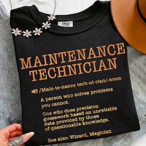 May include: Black t-shirt with the words "MAINTENANCE TECHNICIAN" in orange block letters. Below the title is a definition of the term. A silver necklace with flower accents is visible at the top.