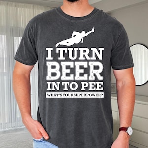 May include: A charcoal gray short-sleeve t-shirt featuring a white graphic of a person holding a bottle. The text reads: I TURN BEER IN TO PEE WHAT'S YOUR SUPERPOWER? The shirt has a faded, vintage-style wash and a classic crew neckline.