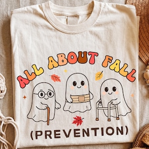 Może przedstawiać: Kremowy t-shirt z grafiką przedstawiającą trzy kreskówkowe duszki. Napis głosi ALL ABOUT FALL (PREVENTION). Jeden duszek nosi okulary i laskę, drugi pasek, a trzeci używa chodzika. Wokół rozrzucone są małe czerwone i żółte liście.