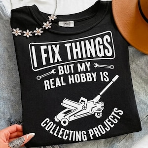 May include: Black crew neck t-shirt with white text that reads "I FIX THINGS BUT MY REAL HOBBY IS COLLECTING PROJECTS". The graphic includes wrenches and a car jack. A floral necklace is visible.