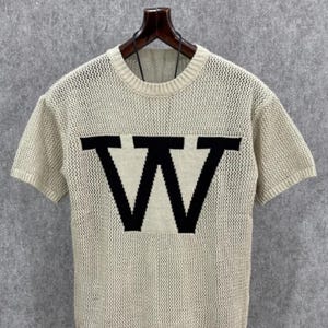 May include: A short-sleeved, cream-colored knit shirt with a large black "W" design across the chest. The shirt has a crew neck and a loose, open weave. The shirt is hanging on a wooden hanger against a gray background.