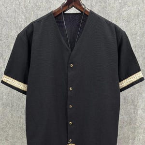May include: Black short-sleeved shirt with a V-neck and button-up front. The sleeves have a decorative cream-colored trim. The shirt is hanging on a wooden hanger against a neutral background. A small piece of burlap is sewn on the bottom.
