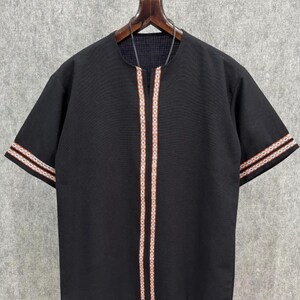 May include: Black short-sleeve shirt with a round neckline and decorative trim. The trim features a pattern of white, orange, and red stripes. The sleeves also have a similar striped design. The shirt is hanging on a wooden hanger.