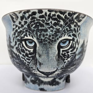 Könnte beinhalten: Keramikschale mit einem Leoparden-Gesichtsmotiv. Die Schale hat einen hellblauen Sockel mit schwarzen Flecken und blauen Augen. Ein einzigartiges Dekorationsstück für den Innenbereich.