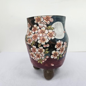 Può includere: Vaso in ceramica dipinto a mano con un motivo floreale. Il vaso presenta uno sfondo blu scuro con una luna piena e fiori di ciliegio nei toni del rosa e del bianco. Il vaso ha tre piccoli piedini arrotondati.