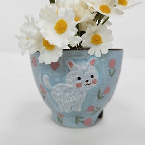 May include: A light blue ceramic planter with a white cat design and pink accents, holding white and yellow daisy flowers. The planter has a rounded shape and small feet.
