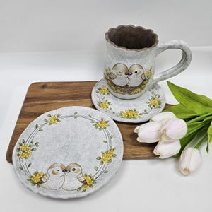 May include: A ceramic teacup, saucer, and plate set. The light grey pieces have a textured finish and feature a design of two kissing birds in a nest, surrounded by yellow flowers. The cup has a large handle and a scalloped rim. The plate is round.