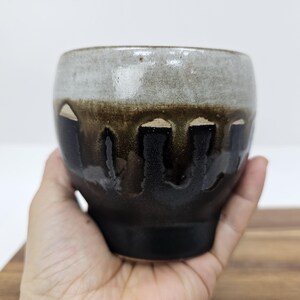 Könnte beinhalten: Eine handgefertigte Keramiktasse mit einer einzigartigen Glasur. Die Tasse weist einen Farbverlauf auf, beginnend mit einem hellgrauen Rand, übergehend in eine dunkelbraune Mitte und endend mit einem schwarzen Boden. Die Glasur hat einen Tropfeffekt, der ihren künstlerischen Reiz verstärkt.
