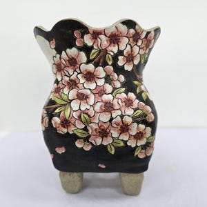 Puede incluir: Una maceta de cerámica negra con un borde festoneado y tres patas pequeñas. La maceta está decorada con un diseño floral de flores de cerezo rosas y blancas y hojas verdes.