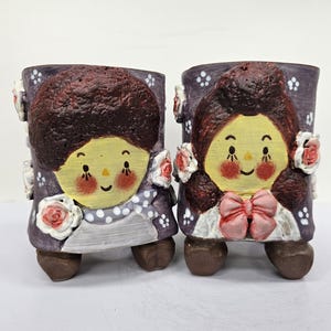 May include: Two ceramic plant pots with cartoon faces. The pots have a purple background and feature a yellow face, brown hair, and a pink bow or collar. White flowers are attached to the sides.