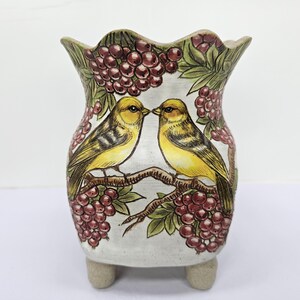Puede incluir: Un jarrón de cerámica con borde festoneado, con un diseño pintado a mano de dos pájaros amarillos posados en una rama con bayas rojas y hojas verdes. El jarrón descansa sobre tres pequeños pies redondeados.