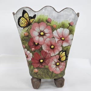 May include: A ceramic planter with a wavy rim, decorated with pink and red flowers, green leaves, and yellow butterflies. The planter stands on three brown feet. The background is light grey. The planter is suitable for indoor use.