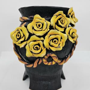 Peut inclure: Vase en céramique noire avec des roses jaunes. Le vase présente une surface texturée et est orné de roses jaunes tridimensionnelles détaillées. Un motif en forme de corde marron entoure le vase. Le vase a une base évasée unique.