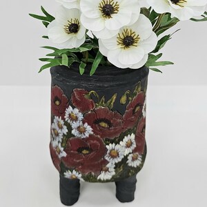 Puede incluir: Un jarrón cilíndrico negro con un diseño floral de amapolas rojas y margaritas blancas. El jarrón tiene tres patas cortas y negras y contiene flores de anémona blancas con centros oscuros y hojas verdes. Una pieza decorativa para el hogar.