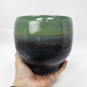 Puede incluir: Un cuenco de cerámica hecho a mano con un degradado de colores. La mitad superior es de un verde vibrante, que pasa a un gris verdoso más oscuro y, finalmente, a un negro intenso en la base. El cuenco tiene forma redondeada y un acabado liso y brillante.