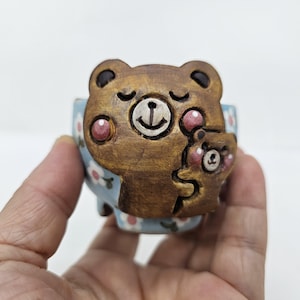 May include: A brown ceramic bear-shaped planter with a smaller bear hugging the larger one. The planter has a light blue background with white and pink floral patterns. The bears have pink cheeks and closed eyes.