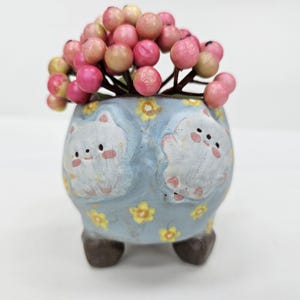 Op de afbeelding: Een kleine, lichtblauwe keramische plantenbak met een grillig ontwerp van twee witte kattengezichten. De plantenbak is versierd met gele bloemen en gevuld met roze en beige bessen. De plantenbak heeft drie bruine voetjes.
