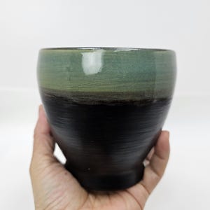 Puede incluir: Un cuenco de cerámica hecho a mano con un esmalte de dos tonos. La mitad superior es verde apagado, que pasa a una banda marrón oscuro y luego a una base negra. El cuenco se sostiene en una mano sobre un fondo blanco.