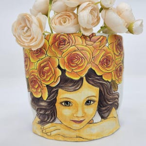 May include: A ceramic flower pot featuring a hand-drawn illustration of a girl with a floral crown. The pot is white with a yellow and orange rose design around the top. The girl's face is a warm yellow color, and her hair is brown.