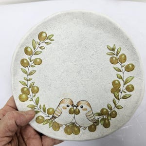 Puede incluir: Un plato redondo de cerámica blanquecina con una superficie texturizada. El plato presenta un diseño pintado de dos pájaros besándose, rodeados por una corona de ramas de olivo y aceitunas en tonos verdes y dorados.