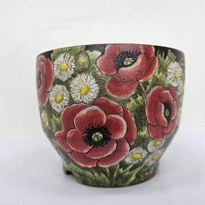 Puede incluir: Jardinera de cerámica pintada a mano con un diseño floral de amapolas rojas y margaritas blancas sobre un fondo verde y negro. La maceta tiene forma redondeada y pequeños pies.