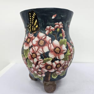 Puede incluir: Jardinera de cerámica decorativa con fondo azul oscuro, adornada con flores rosas y blancas, hojas verdes y una mariposa amarilla y negra. La jardinera tiene tres patas marrones.