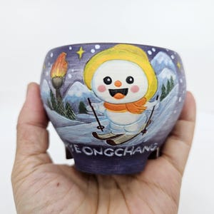 May include: A hand-painted ceramic pot featuring a snowman skiing down a snowy mountain. The pot is decorated with a torch, mountains, and the word "JEONGCHANG". The snowman wears a yellow hat and orange scarf.