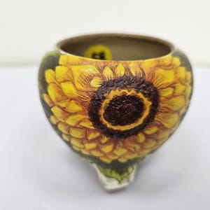 May include: A ceramic container with a sunflower design. The container is painted with yellow, brown, and green colors. The sunflower design covers the exterior of the container, with a detailed depiction of the flower's petals and center.