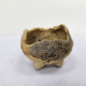 Może przedstawiać: Mały, ręcznie robiony ceramiczny garnek o teksturowanym, ziemistym wyglądzie. Garnek jest beżowy z ciemnobrązowymi i czarnymi akcentami i spoczywa na trzech małych nóżkach. Obręcz ma nieregularny, organiczny kształt.