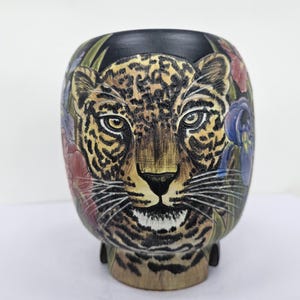 Könnte beinhalten: Dekorative Keramikvase mit detailliertem Leoparden-Gesicht-Design. Die Vase hat einen schwarzen Rand und Boden mit bunten floralen Akzenten. Das Gesicht des Leoparden ist in Braun-, Beige- und Schwarztönen gehalten.